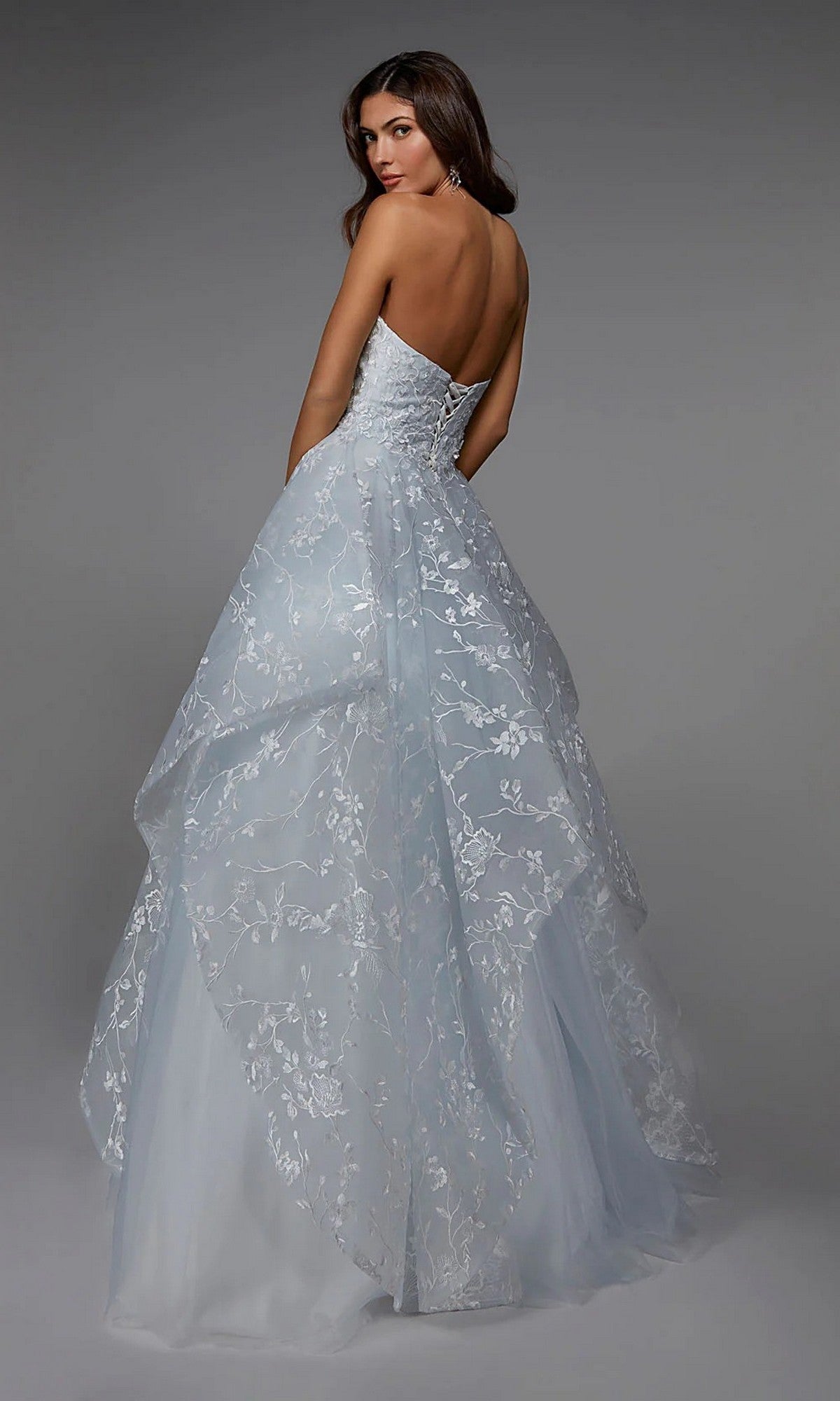 Woman in the Alyce 7041 long formal dress, a strapless and backless ball gown with layered sheer fabric and floral embroidery, stands against a plain gray backdrop, glancing over her shoulder.